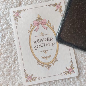 Könnte beinhalten: Eine rechteckige Karte mit cremefarbenem Hintergrund, einem goldenen ovalen Rahmen, einer rosa Schleife und floralen Akzenten in den Ecken. Der Text "READER SOCIETY" steht in der Mitte. Eine schwarze, glitzernde Smartphone-Hülle verdeckt die Karte.
