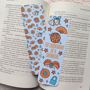 May include: Two light blue bookmarks with orange and white illustrations of hearts, fans, globes, and stars. One bookmark reads "THE BOOKMARK OF A SHOWGIRL". The bookmarks are placed on an open book.