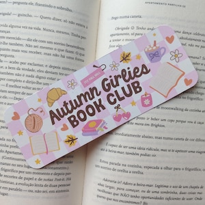 Pode incluir: Um marcador de livros colorido com o texto "Autumn Girlies BOOK CLUB" e várias ilustrações com tema de outono. O marcador de livros apresenta um padrão xadrez rosa e branco, com imagens de um croissant, cerejas e uma xícara de café.