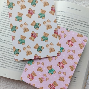 Op de afbeelding: Twee rechthoekige kaarten met een herhaald patroon van bruine teddyberen met truien en hoeden. Eén kaart is wit, de andere is roze. De kaarten liggen naast een open boek.