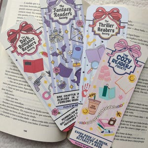 May include: Four colorful bookmarks with different themes: "The Dark Romance Readers Society", "The Fantasy Readers Society", "The Thriller Readers Society", and "The Cozy Readers Society". Each bookmark has unique illustrations and text, including "Every book is a mood, and every mood deserves a book."