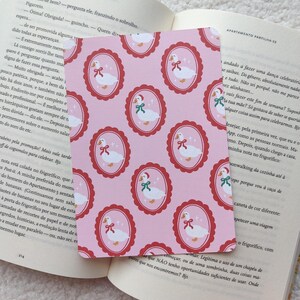 Op de afbeelding: Roze kaart met een herhaald patroon van witte ganzen in ovale kaders met rode randen en strikken. De kaart ligt op een open boek.