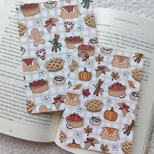 Könnte beinhalten: Zwei dekorative Lesezeichen mit einem sich wiederholenden Muster von herbstlichen Illustrationen, darunter Kuchen, Kürbisse und Zimtstangen, auf einem beige-weiß karierten Hintergrund. Die Lesezeichen liegen auf einem offenen Buch.