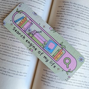 Pode incluir: Um marcador de página retangular com fundo verde claro apresentando um gráfico de barra de pesquisa rosa contendo livros coloridos e um ícone de lupa. O texto "Search engine of my life" está impresso em preto, com pequenos detalhes florais.