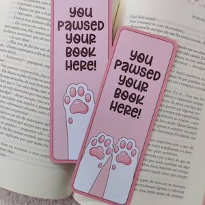 Può includere: Due segnalibri rettangolari rosa con angoli arrotondati. Ogni segnalibro presenta il testo marrone scuro 'you paused your book here!' sopra una zampa di gatto bianca in stile cartoon con cuscinetti rosa, appoggiati su un libro aperto.