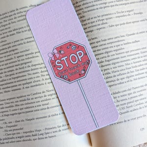 Może przedstawiać: Jasnofioletowa prostokątna zakładka do książki z grafiką czerwonego ośmiokątnego znaku stopu, różową kokardką, małymi ilustracjami truskawek oraz napisem "STOP don't touch my book" w kolorze białym i czarnym, leżąca na otwartej stronie.