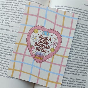 Peut inclure: Un marque-page en forme de cœur rose et blanc avec le texte "Just A Silly Bookish Girlie" et des illustrations. Il est posé sur un livre avec un fond quadrillé.