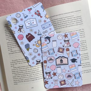Pode incluir: Dois marcadores de livros azuis claros com ilustrações com tema de praia, incluindo um urso de óculos de sol, conchas e o texto "Entering Cousins Beach". Os marcadores de livros estão em um livro aberto.