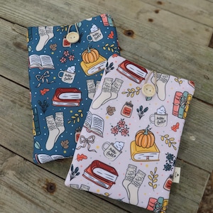 Pode incluir: Duas capas de livros com padrões de outono. Uma é azul com livros, meias, abóboras e canecas com a frase "Hello Fall". A outra é rosa claro com motivos semelhantes. Ambas têm fechos de botão de madeira.