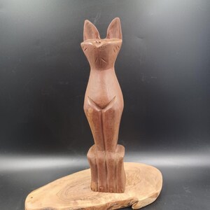 May include: A wooden sculpture of a cat standing on a wooden base. The cat is brown and has a simple, stylized design.