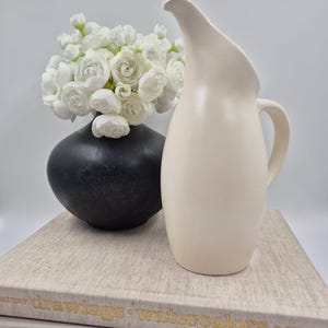 May include: A matte cream-colored ceramic pitcher with a curved spout and handle, positioned next to a black spherical vase filled with white flowers, all resting on two stacked books titled drawings: watercolors and IS MON AMOUR.