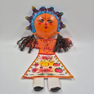 May include: A handmade folk art doll with an orange face, blue and yellow sun-like halo, and black yarn hair. The doll wears an orange dress with a fruit-decorated apron and white legs with black shoes. A unique, colorful piece.