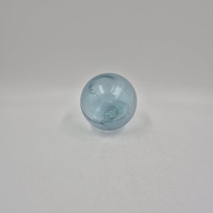 May include: A clear, light blue glass ball. The sphere has a slightly irregular shape and internal swirls, suggesting it may be handmade. The glass has a subtle, translucent quality, allowing light to pass through.