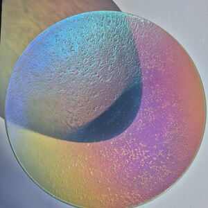May include: A round, iridescent glass plate with a textured surface. The plate reflects a spectrum of colors, including blue, purple, and yellow. A shadow of a similar shape is cast on the white surface behind the plate.