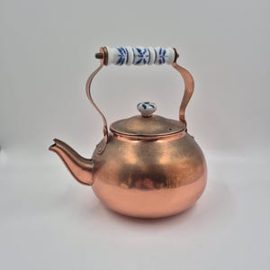 May include: A copper tea kettle with a rounded body and a curved spout. The lid features a small white ceramic knob with blue floral patterns. The handle is made of white ceramic segments decorated with blue floral designs and held by copper arms.