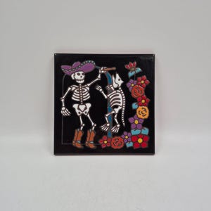 May include: A square black ceramic tile featuring a colorful graphic of a skeleton wearing a purple sombrero and brown boots, standing next to a skeleton dog. The design includes a blue archway and a vertical arrangement of red, orange, and purple flowers.