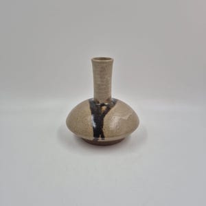 May include: A handcrafted ceramic vase with a unique shape. The vase features a light brown body with a dark brown vertical stripe and a narrow, cylindrical neck. The vase is a decorative item.