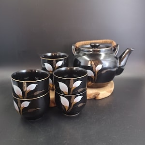 May include: A black tea set with a teapot and four cups. The teapot and cups are decorated with white flowers and gold accents.