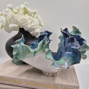 May include: A ceramic sculpture with a white base and ruffled, wavy edges featuring a mottled glaze in shades of deep blue, teal, and hints of purple. The piece has a fluid, organic shape with a glossy finish and a textured, uneven rim.