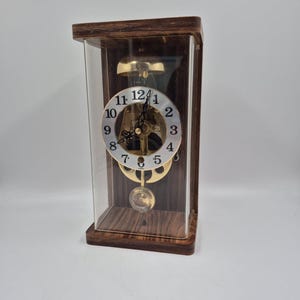 May include: A rectangular mantel clock with a dark wood frame and a clear glass enclosure. The clock features a circular white face with black Arabic numerals, black hands, and a visible gold-toned mechanical movement with a swinging pendulum.