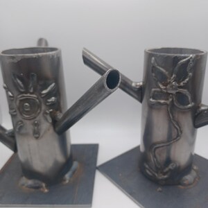 May include: Two metal candle holders with a square base. One holder features a sun design and the other features a flower design.