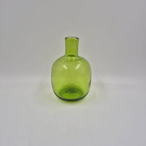 May include: A small, round, green glass bottle with a narrow neck. The bottle has a smooth, clear surface and a slightly rounded base. The green color is consistent throughout the glass, giving it a vibrant appearance. The bottle is empty and stands upright.