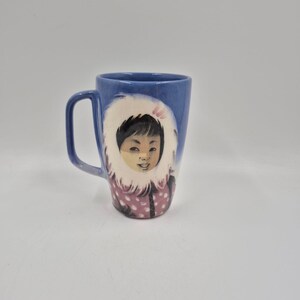 May include: Blue ceramic mug featuring a painted illustration of a child in a winter coat with a white fur-trimmed hood. The mug has a curved handle and a glossy finish, perfect for hot beverages.