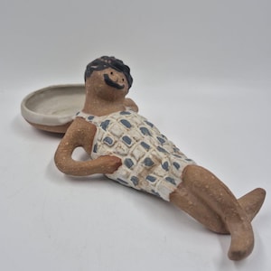 May include: A ceramic figurine of a person reclining, featuring a textured brown clay body, black hair, and a white swimsuit with a blue grid pattern. The figure is positioned with one arm resting on a small, attached circular white bowl.