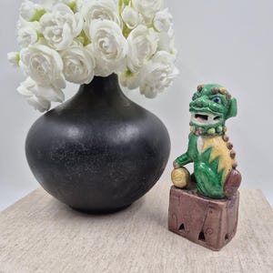 May include: A matte black ceramic vase filled with white flowers next to a green, yellow, and brown glazed ceramic foo dog figurine. The foo dog is perched on a rectangular base and holds a small golden sphere under its right front paw.