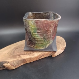 May include: A square ceramic planter with a dark brown, black, and green glaze. The planter has a textured surface and is sitting on a wooden surface.