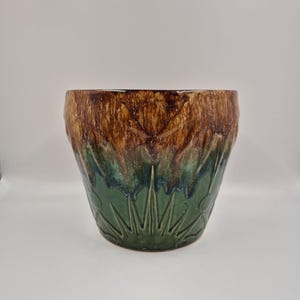 May include: A ceramic plant pot with a textured surface. The top portion is brown, transitioning to green with a leaf-like pattern at the bottom. The pot has a wide, open top and a slightly tapered shape.