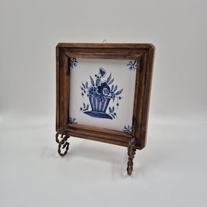 May include: A framed ceramic tile featuring a blue floral design in a vase. The tile is set within a dark wooden frame and supported by decorative metal legs. The artwork is displayed on a white background.