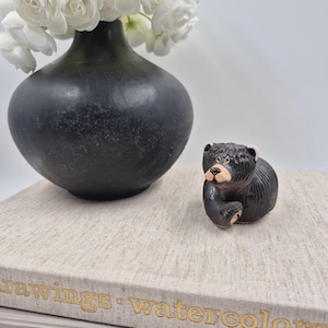 May include: A small, black ceramic bear figurine with a textured coat, a light-colored muzzle, and small ears. The bear is positioned next to a matte black vase filled with white flowers, all resting on a stack of two hardcover books.