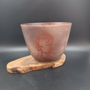 May include: A brown ceramic bowl with a red stylized ram design. The bowl is sitting on a wooden base.