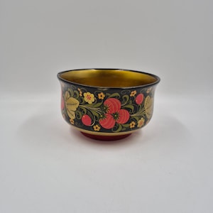 May include: A small, round bowl with a black exterior featuring a hand-painted floral pattern in red, yellow, and green. The interior of the bowl is finished in a metallic gold color, and the base of the bowl is a solid red color.