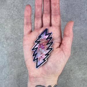 Puede incluir: Un parche decorativo con forma de rayo. El parche presenta un diseño de galaxia colorida con tonos de rosa, morado y azul. El rayo está delineado en negro.