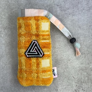 May include: A yellow and cream-colored phone case with a textured waffle pattern. The case features a black and white patch with a geometric design. A patterned strap with a black bead is attached to the case.