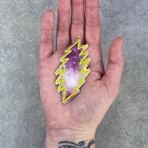 May include: A colorful lightning bolt patch with a galaxy design in purple, pink, and white, outlined in bright yellow. The patch is held in an open hand against a gray background. The patch is approximately 2 inches in length.