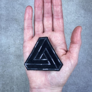 May include: Black patch featuring a Penrose triangle design. The patch is a dark color with a lighter gray outline and internal lines, creating an optical illusion. The patch is held in a hand, showcasing its size and detail.
