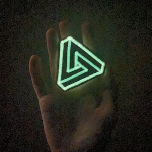 May include: A glowing, triangular pendant with a three-dimensional design, held against a dark background. The pendant emits a bright green light, highlighting its intricate, interlocking lines. The overall effect is a striking, geometric design.