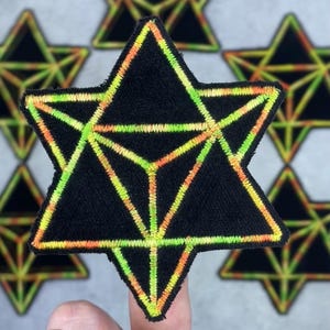 May include: Black star-shaped patch with a geometric design. The patch features a six-pointed star with a three-dimensional design inside. The edges and interior lines are embroidered with neon green and orange thread.