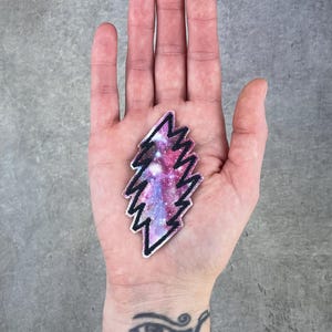 Puede incluir: Un parche en forma de rayo con un diseño de galaxia en tonos rosa, morado y azul. El parche tiene un borde negro y se sostiene en una mano.