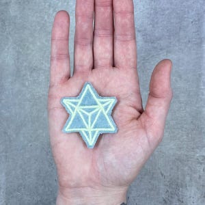 May include: A six-pointed star patch featuring a light blue fabric base with a pale yellow geometric line design embroidered on the surface. The star shape is formed by two overlapping triangles, creating a central triangular pattern.