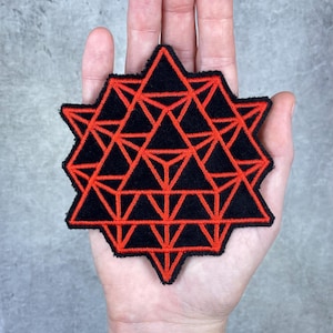 May include: Black and red geometric patch featuring a complex star-like design. The patch has a black background with a red geometric pattern. The patch is held in a hand.