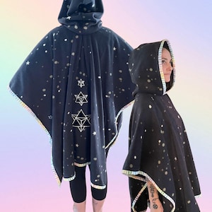 May include: A dark charcoal hooded poncho featuring a scattered white star pattern and a rainbow-striped trim along the edges. The back displays three stacked geometric star symbols in white line art, while the front shows a simple hooded design.