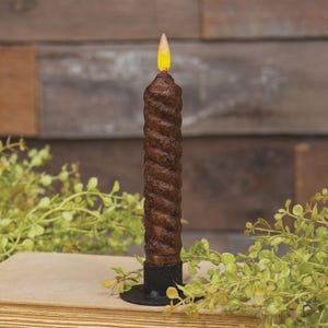 May include: A dark brown, spiral-textured taper candle with a rough, matte finish. The candle is topped with a small, glowing yellow flame-shaped bulb and is secured in a simple, circular black metal candle holder base.