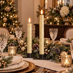 May include: Two tall, cream-colored taper candles with a speckled texture, each lit and standing in a black metal base. They are positioned as a centerpiece among evergreen branches, pinecones, gold ornaments, and small glass votive candle holders.