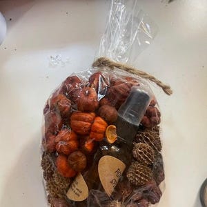 May include: A clear plastic bag tied with twine containing a collection of small orange decorative pumpkins and brown dried seed pods. Inside the bag is a dark glass spray bottle with a black nozzle and a tan label with the text Pumpkin Spice.