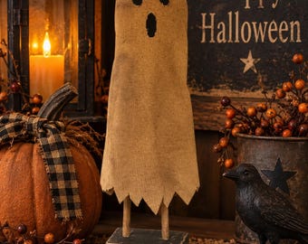 May include: A standing ghost decoration made of tan burlap fabric with two dark circular eyes and an oval mouth, mounted on two wooden dowels attached to a rectangular black base. The ghost has a jagged, uneven bottom edge.