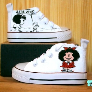 Puede incluir: Zapatillas altas blancas con ribetes negros e ilustraciones de dibujos animados. Una zapatilla presenta un personaje con la palabra "LIBERTAD" en un bocadillo. La otra zapatilla muestra a una niña con un vestido rojo.